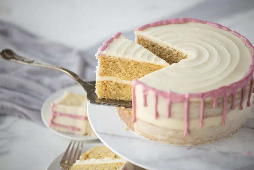 Glutenfreie Vanille-Torte, zweilagig, mit rosa Drip-Glasur und angeschnittener Kuchenscheibe auf Tortenheber