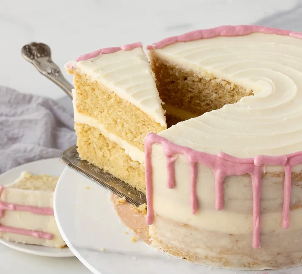 Zweilagige glutenfreie Vanille-Torte mit rosa Drip-Dekor und angeschnittenem Stück auf Tortenplatte