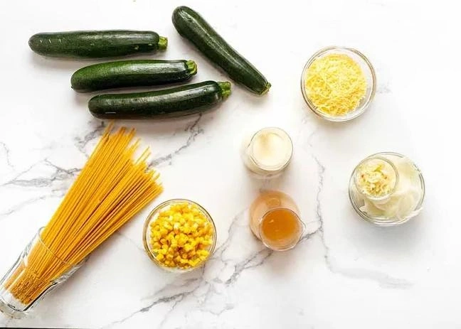 Zutaten für Spaghetti mit Zucchinisauce: Zucchini, Pasta, Mais, Sahne, Parmesan, Zwiebeln, Knoblauch und Brühe auf hellem Marmorhintergrund.