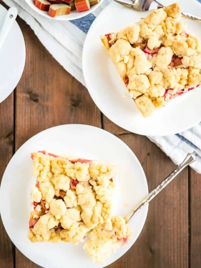 Zwei Stück Omas Rhabarber Streuselkuchen vom Blech auf weißen Tellern mit Löffel, serviert auf rustikalem Holztisch.
