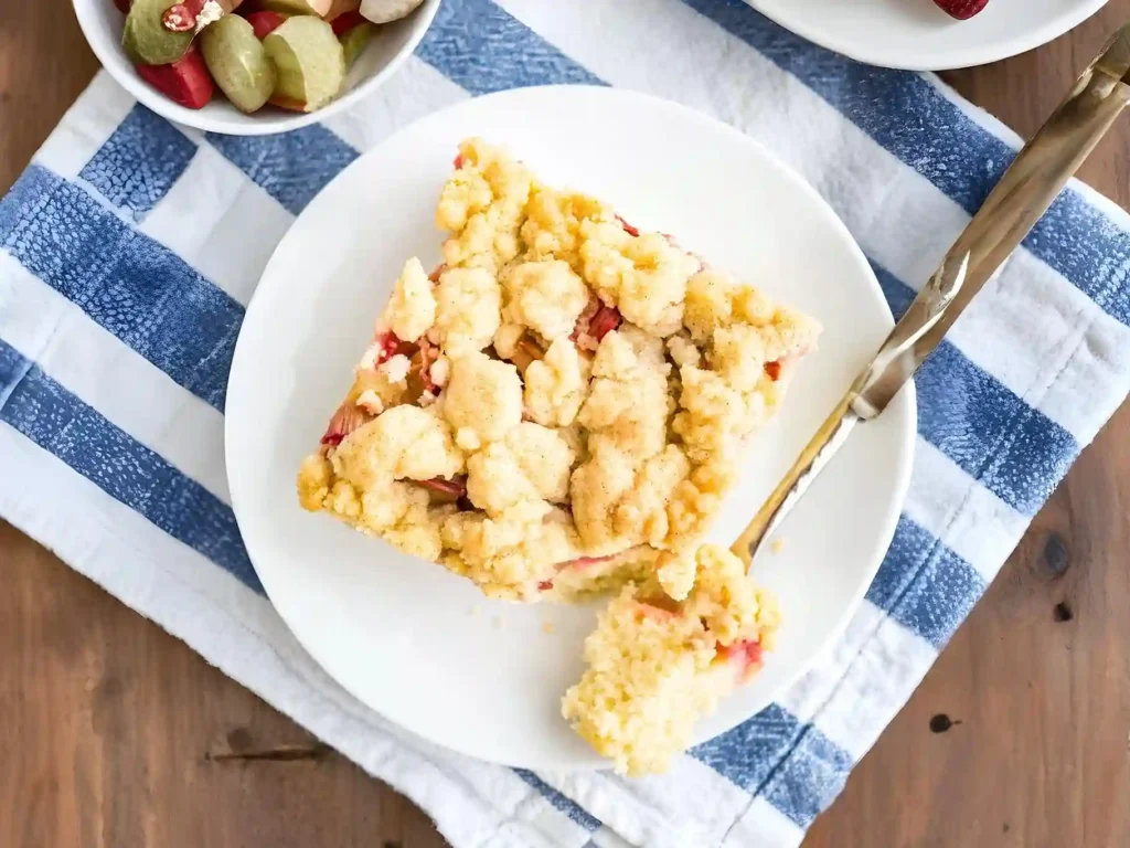 Omas Rhabarber Streuselkuchen vom Blech mit goldbraunen Streuseln auf weißem Teller, serviert mit Löffel und frischem Rhabarber.