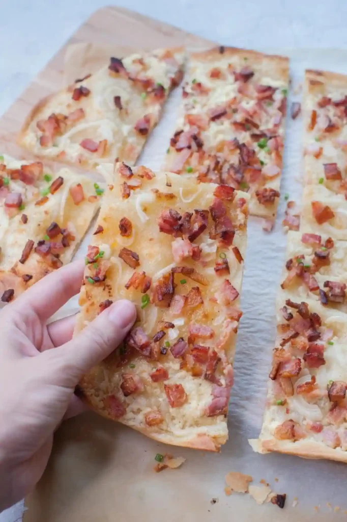 Hand hält ein Stück Elsässer Flammkuchen mit Speck und Zwiebeln
