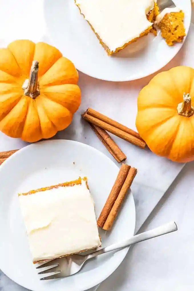 Kürbiskuchen vom Blech mit Frischkäse Frosting auf Tellern mit Zimtstangen und Kürbissen