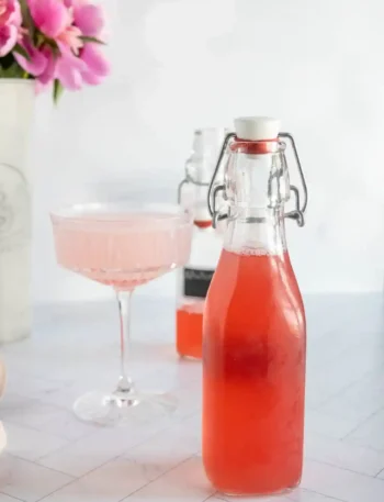 Rhabarbersirup selber machen – fruchtiger Rhabarber Sirup in Glasflasche mit Cocktailglas im Hintergrund