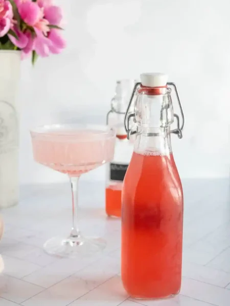 Rhabarbersirup selber machen – fruchtiger Rhabarber Sirup in Glasflasche mit Cocktailglas im Hintergrund