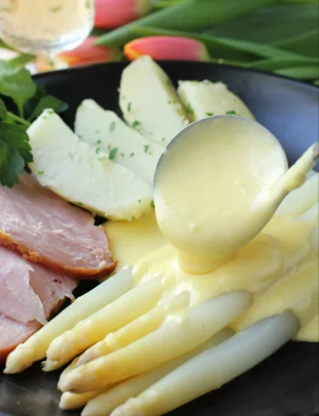 Spargel mit Sauce Hollandaise auf dem Teller – weiße Spargelstangen mit cremiger Soße zu Spargel, Kartoffeln und Schinken
