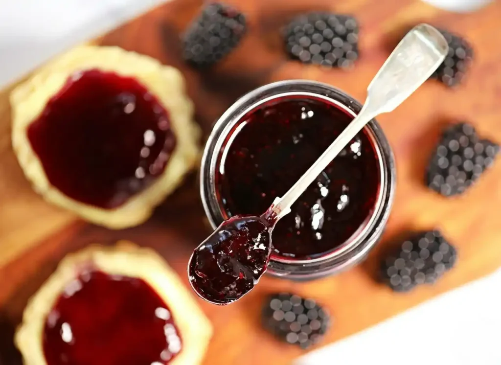 Brombeermarmelade ohne Kerne im offenen Glas mit Löffel, frischen Brombeeren und bestrichenen Scones.