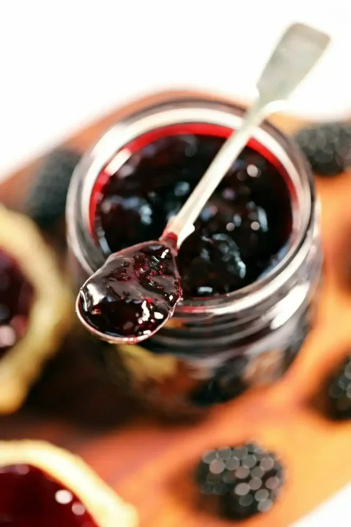 Löffel mit selbst gemachter Brombeermarmelade ohne Kerne über einem offenen Marmeladenglas mit frischen Brombeeren.