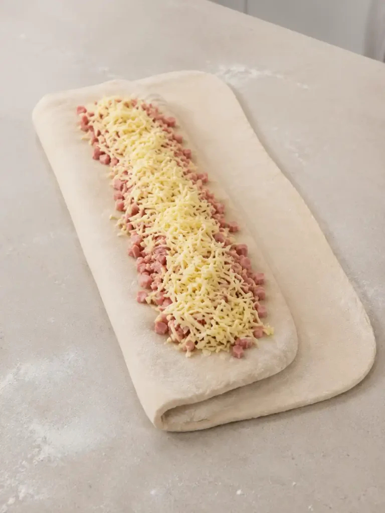 Teilweise gefalteter Handbrot-Teig mit einer zweiten Füllung aus Schinkenwürfeln und geriebenem Käse.