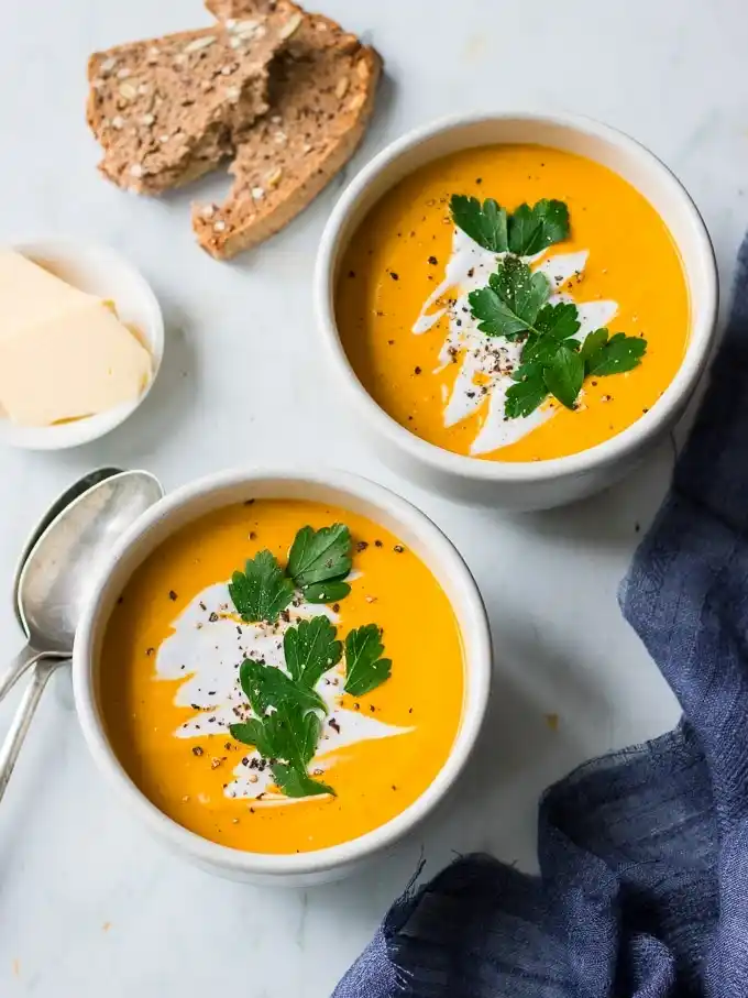 Zwei Schalen cremige Hokkaido-Kürbissuppe mit Kokosmilch und Petersilie, serviert mit Vollkornbrot und Butter.