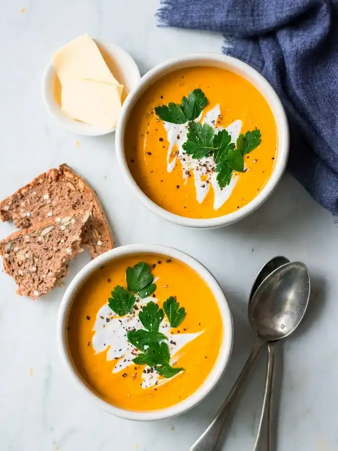 Draufsicht auf zwei Schalen Hokkaido-Kürbissuppe mit Kokosmilch und Petersilie, serviert mit Körnerbrot und Butter.