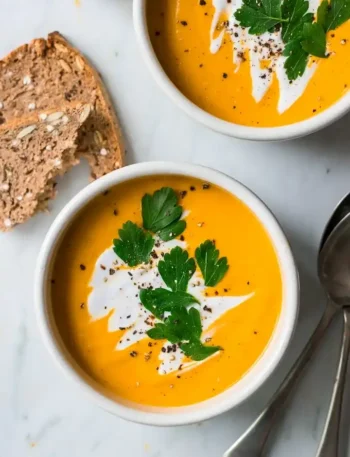 Cremige Hokkaido-Kürbissuppe mit Kokosmilch, frischer Petersilie und Vollkornbrot in zwei weißen Schalen.