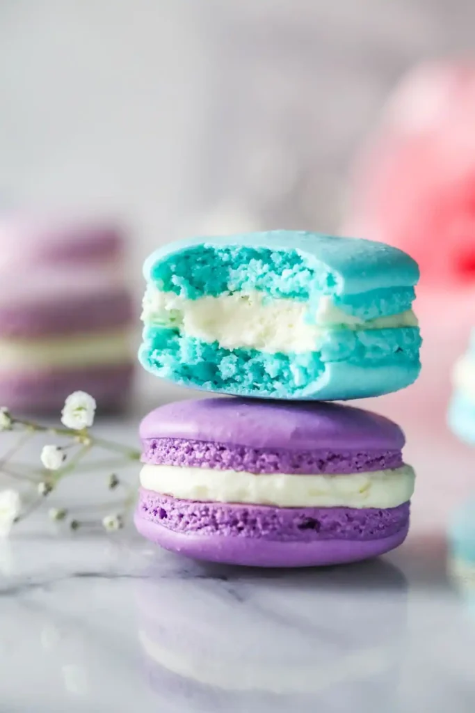 Angebissenes türkises Macaron auf einem lilafarbenen französischen Macaron mit heller Cremefüllung und typischen Füßchen.
