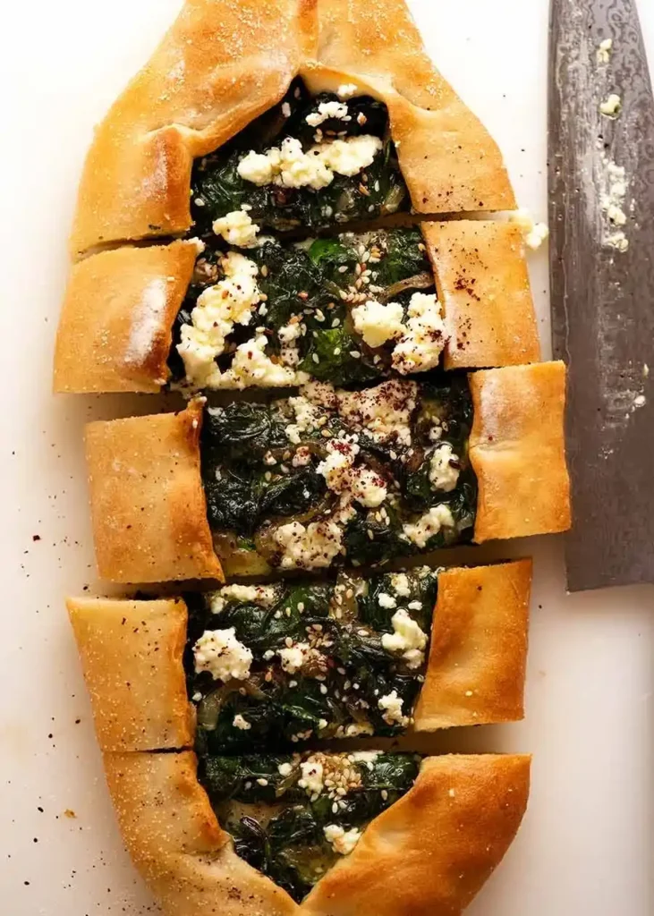 Aufgeschnittene türkische Pide mit Spinat, Feta, Sesam und Sumach auf einem hellen Brett.