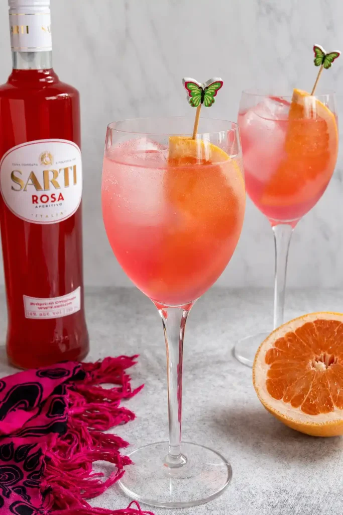 Nahaufnahme eines pinken Sarti Spritz mit Sarti Rosa, Prosecco, Soda, Eiswürfeln und Grapefruit, dahinter ein zweites Glas und die Flasche.
