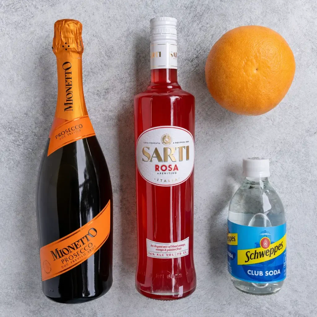 Zutaten für Sarti Spritz: Sarti Rosa, Prosecco, Soda und Grapefruit auf einer hellen Arbeitsfläche.