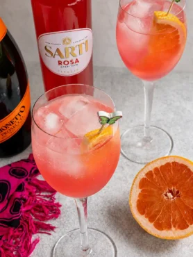 Zwei Gläser Sarti Spritz mit Sarti Rosa, Prosecco, Soda, Eiswürfeln und Grapefruit neben den passenden Flaschen.
