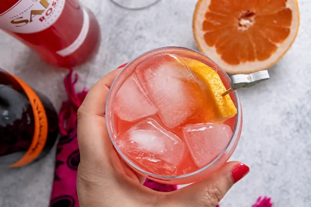 Hand hält einen pinken Sarti Spritz mit Sarti Rosa, Prosecco, Soda, großen Eiswürfeln und Grapefruitgarnitur.