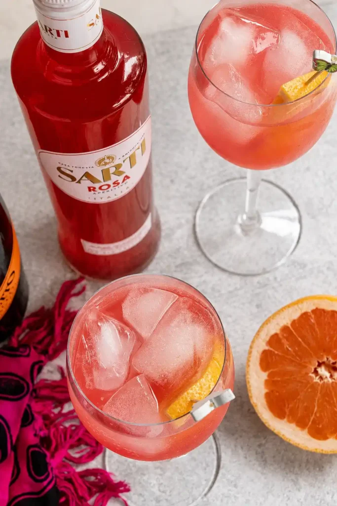 Draufsicht auf zwei Gläser Sarti Spritz mit Eis und Grapefruit neben Sarti Rosa, Prosecco und einer halben Grapefruit.