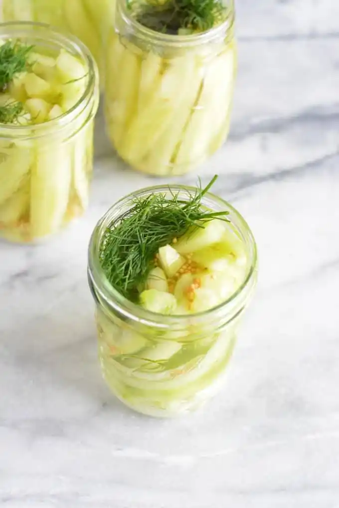 Drei offene Gläser mit selbst eingelegten Senfgurken, frischem Dill und Senfkörnern auf einer hellen Marmorfläche.
