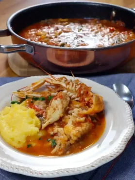 Brudet aus Dalmatien mit Fischstücken, Kaisergranat und Polenta auf einem weißen Teller, dahinter ein Topf mit Fischeintopf.