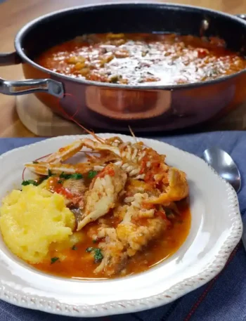 Brudet aus Dalmatien mit Fischstücken, Kaisergranat und Polenta auf einem weißen Teller, dahinter ein Topf mit Fischeintopf.