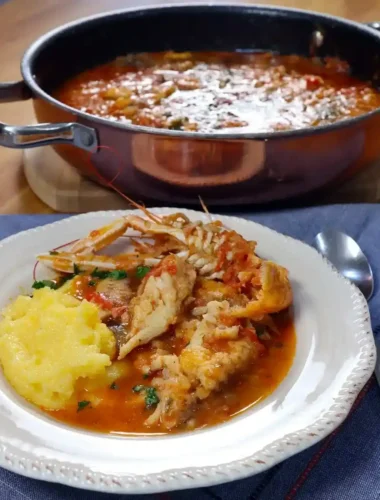 Brudet aus Dalmatien mit Fischstücken, Kaisergranat und Polenta auf einem weißen Teller, dahinter ein Topf mit Fischeintopf.