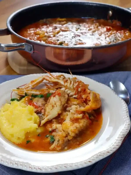 Brudet aus Dalmatien mit Fischstücken, Kaisergranat und Polenta auf einem weißen Teller, dahinter ein Topf mit Fischeintopf.