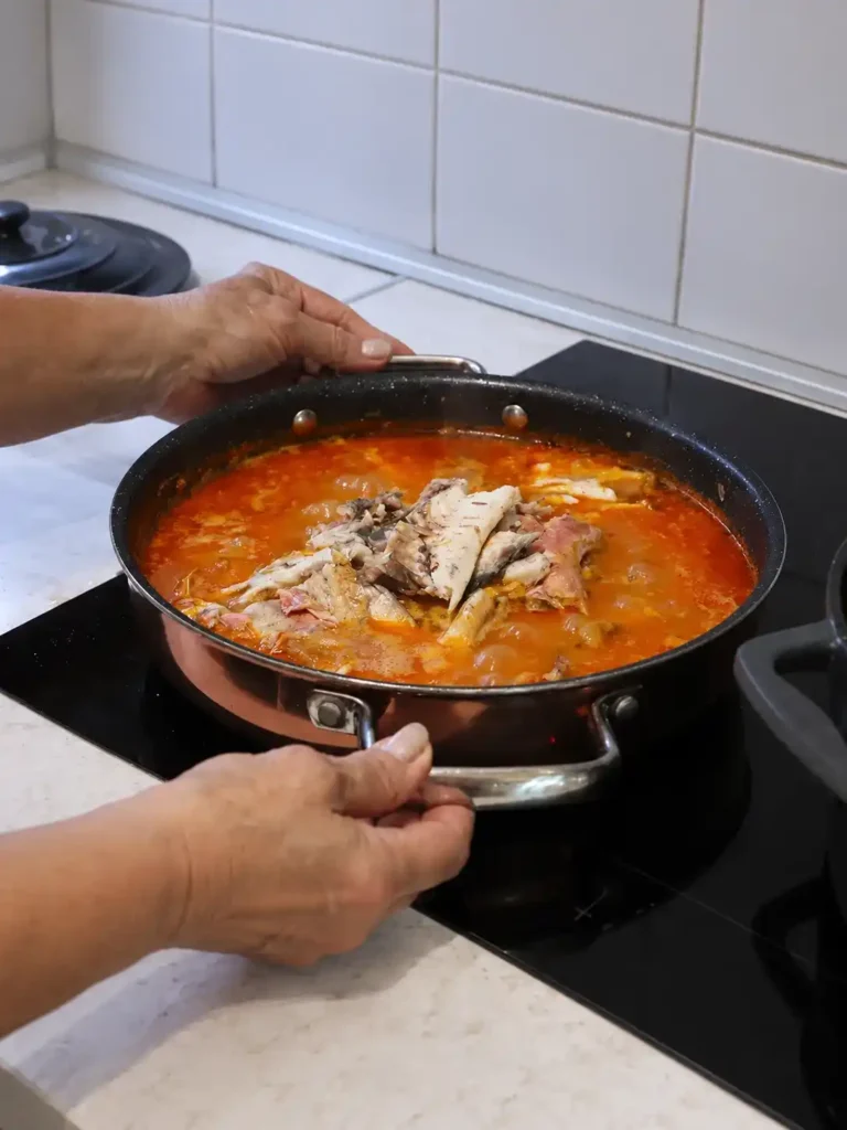 Hände halten die Griffe einer Pfanne mit Brudet, Fischstücken und roter Tomatensoße auf einem schwarzen Kochfeld.