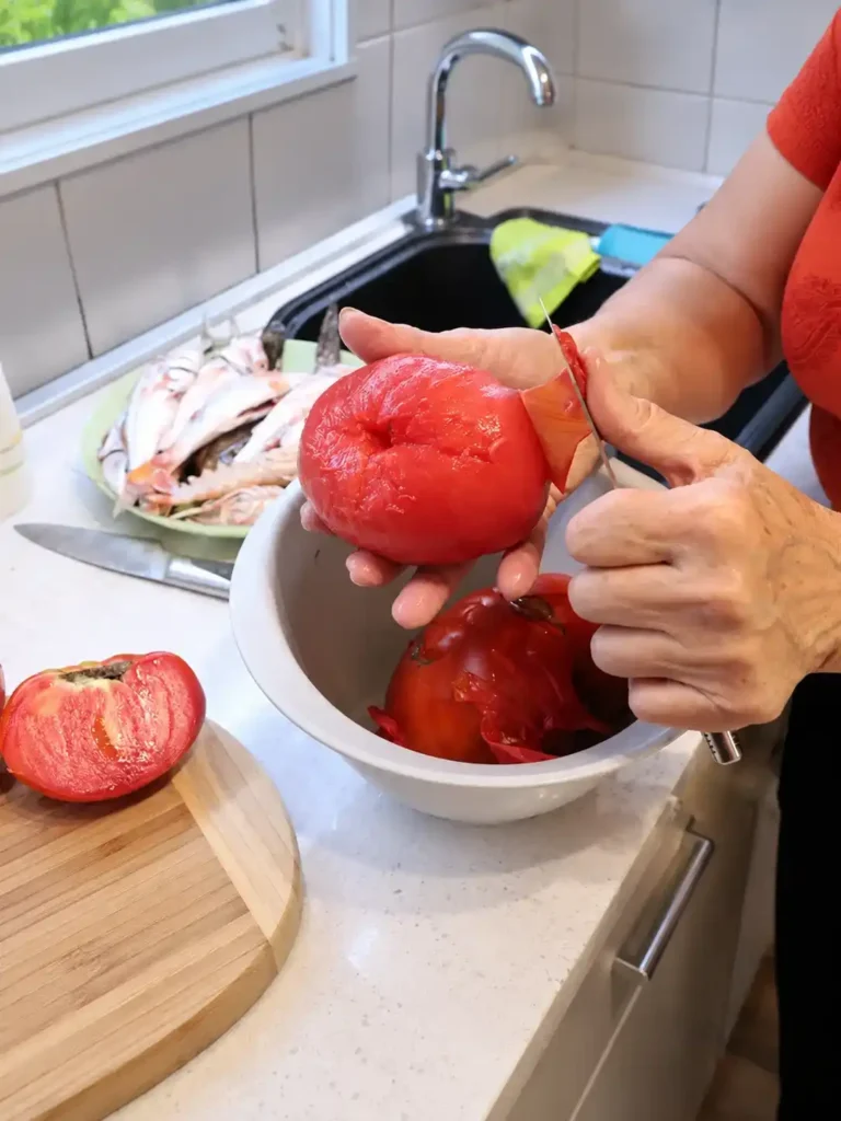 Eine Tomate wird für das Brudet-Rezept mit einem Messer über einer Schüssel gehäutet, im Hintergrund liegen ganze Fische.