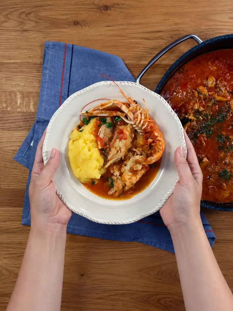 Hände halten einen weißen Teller mit Brudet, Fischstücken, Kaisergranat und Polenta neben dem Topf mit Fischeintopf.
