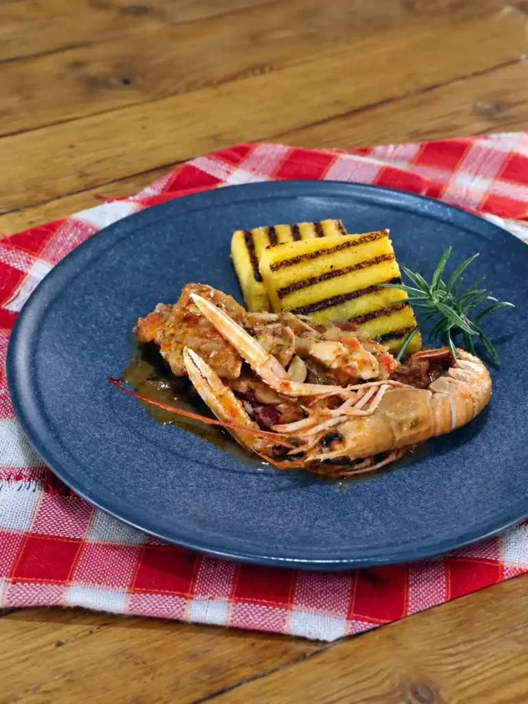 Brudet mit Fischstücken, Kaisergranat und gegrillten Polentaschnitten auf einem blauen Teller mit Rosmarinzweig.