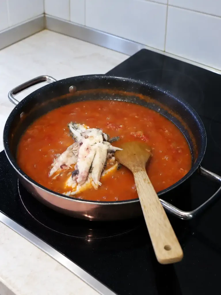 Fischstücke für Brudet in roter Tomatensoße in einer breiten Pfanne auf dem Herd, daneben ein Holzspatel.