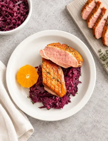 Entenbrust mit goldbrauner Haut und rosa Anschnitt auf Rotkohl, mit einer Orangenscheibe auf einem weißen Teller.