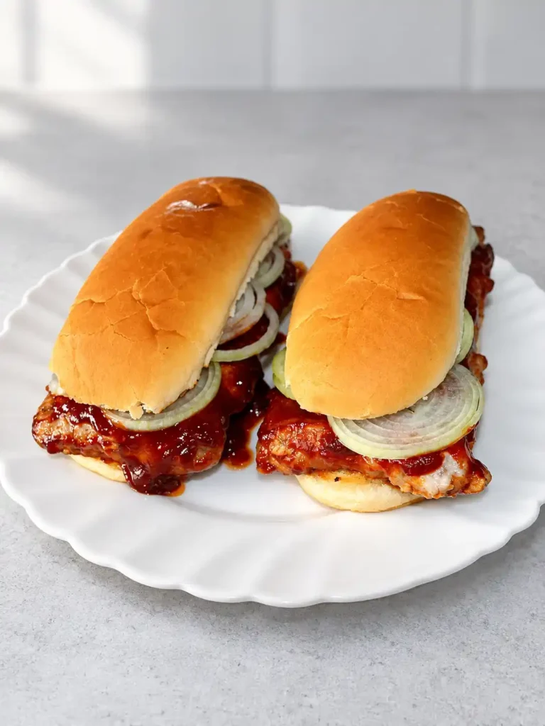 Zwei McRib-Sandwiches mit BBQ-Soße, Zwiebelringen und Gewürzgurken in goldbraunen Brötchen auf einem weißen Teller.