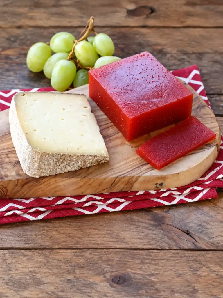 Rotes Quittenbrot mit einer abgeschnittenen Scheibe, Käse und grünen Trauben auf einem Holzbrett.