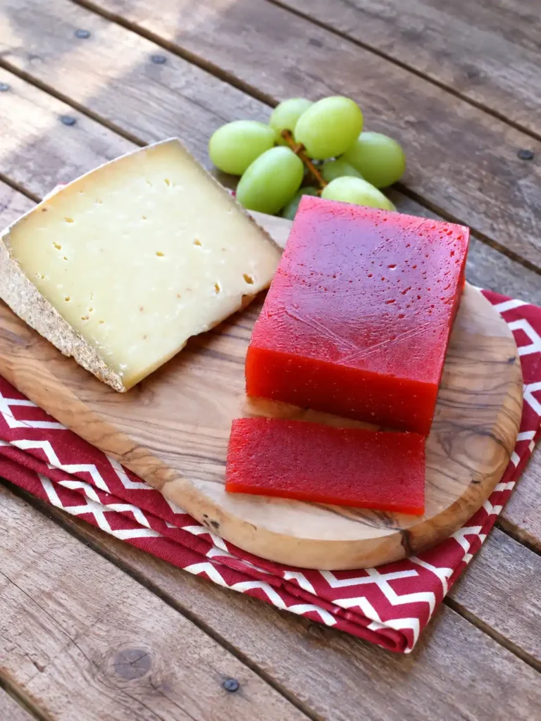 Quittenbrot als roter Block mit einer dünnen Scheibe, Käse und grünen Trauben, schräg von oben fotografiert.