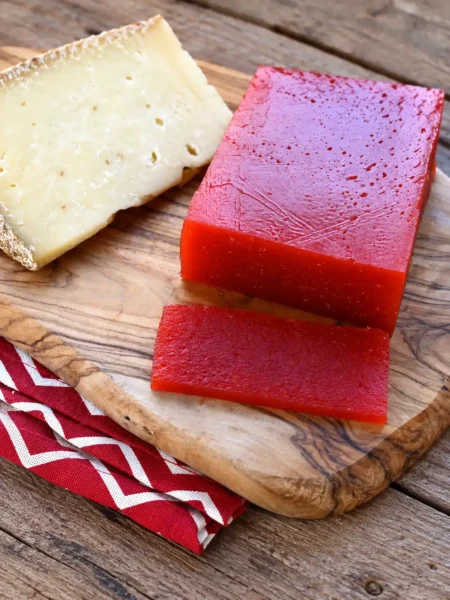 Rotes Quittenbrot als Block und Scheibe neben einem Stück Käse auf einem Holzbrett.