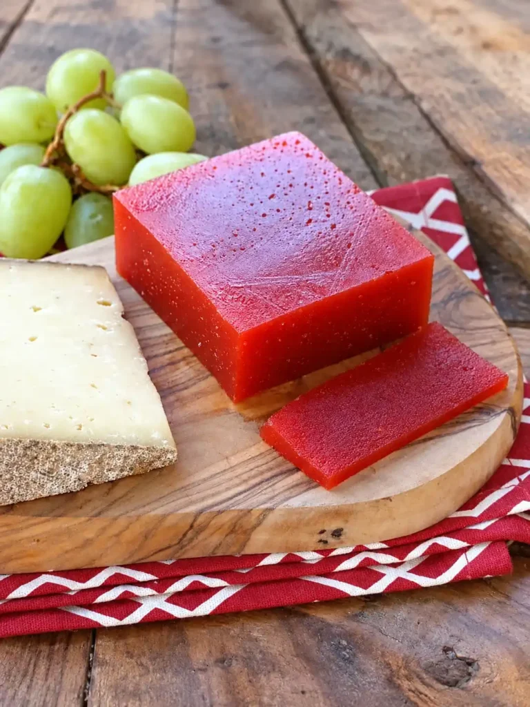 Rotes Quittenbrot mit abgeschnittener Scheibe, Käse und grünen Trauben auf einem Holzbrett.